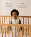 A toddler standing inside a light wooden cot wearing an animal-patterned 1.0 TOG Sleeping Bag, accompanied by product feature callouts for 