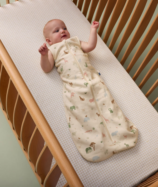 Baby lying in a cot, wearing a cream-colored sleeping bag with safari animal print