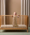 A smiling baby stands inside a wooden cot, holding onto the railing with one hand and a small plush toy in the other. The baby is dressed in a sleeveless cream-colored sleeping bag featuring a colorful safari animal print.
