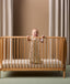 A smiling baby stands inside a wooden cot, holding onto the railing with one hand and a small plush toy in the other. The baby is dressed in a sleeveless cream-colored sleeping bag featuring a colorful safari animal print.