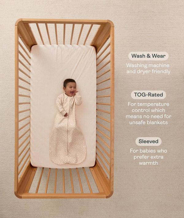 An overhead view on an infant lying in a wooden cot with a grid-patterned sheet, wearing a long sleeve Sleeping Bag 1.0 TOG with text highlighting 