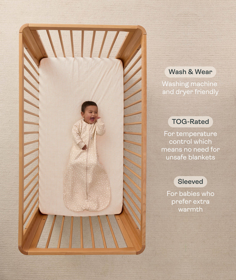 An overhead view on an infant lying in a wooden cot with a grid-patterned sheet, wearing a long sleeve Sleeping Bag 1.0 TOG with text highlighting 