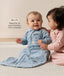 Two babies wearing long-sleeved sleeping bags sit on a carpet, with text highlighting slim-fit design for an easy transition from swaddling.