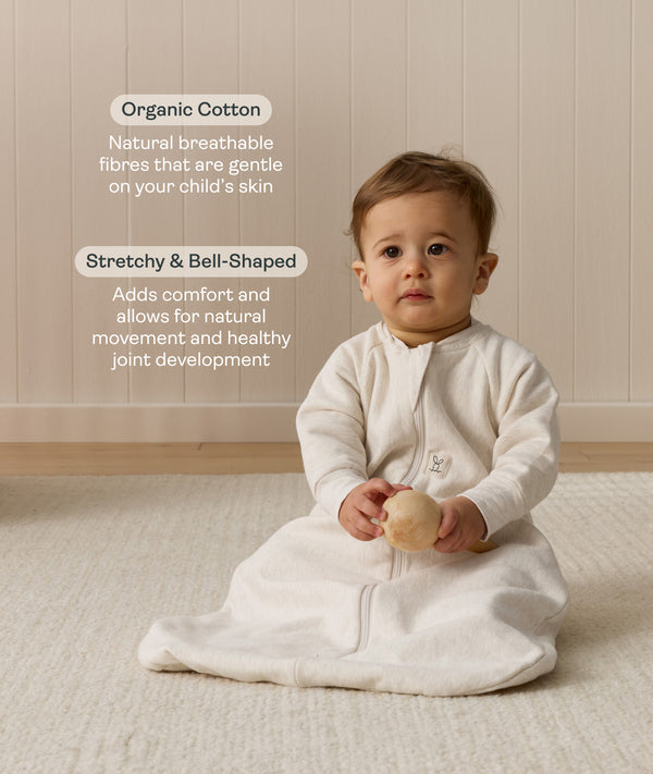 Infant sitting, wearing a sleeved Sleeping Bag 1.0 TOG Oatmeal Marle, while holding a wooden toy. Text highlighting organic cotton and stretchy-bell-shaped design.
