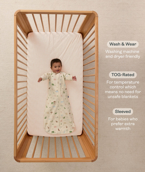 Overhead view of a baby lying on their back in a wooden cot, wearing a safari-patterned long-sleeve Sleeping Bag with text highlighting wash & wear, TOG rating, and sleeve features.