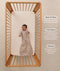 A happy baby lying in a wooden cot, wearing a sleeved Sleeping Bag 1.0 TOG Taupe Marle with text highlighting 