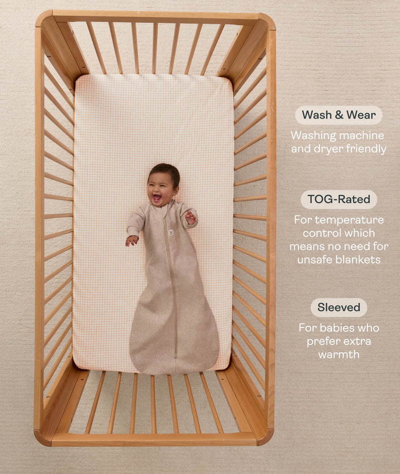 A happy baby lying in a wooden cot, wearing a sleeved Sleeping Bag 1.0 TOG Taupe Marle with text highlighting 