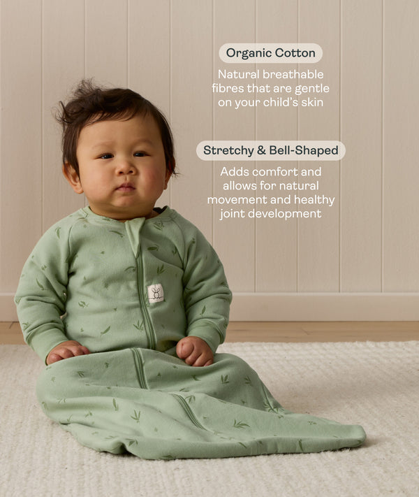 Infant sitting on a white mat, wearing a Sleeved Sleeping Bag 1.0 TOG Willow with text overlay highlighting organic cotton and stretchy & bell-shaped design.