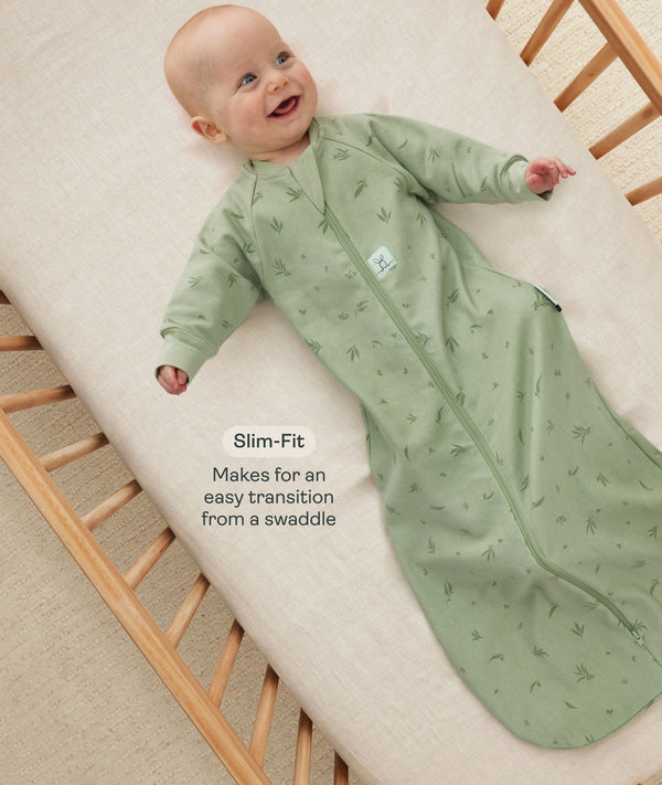 Happy baby lying in a wooden cot wearing a green long-sleeved sleeping bag with text overlay highlighting slim-fit design for an easy transition from swaddling.