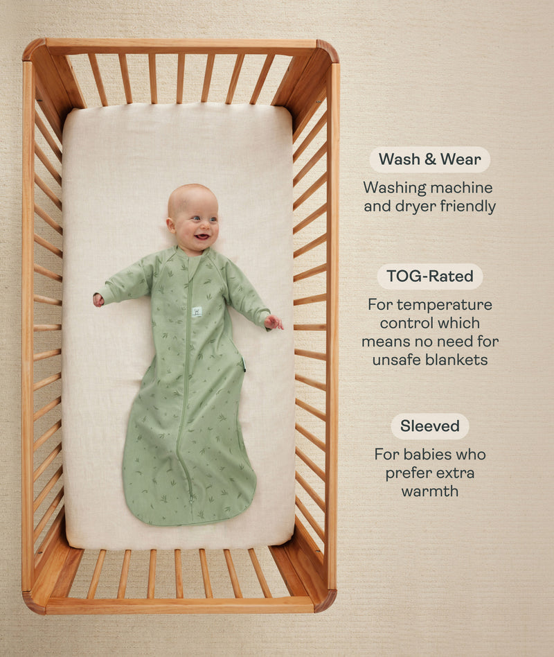 An overhead view of a baby wearing a long-sleeved, sage green leaf-patterned sleeping bag lying on their back inside a wooden cot. To the right, text details key features: wash & wear, TOG-rated, and Sleeved.