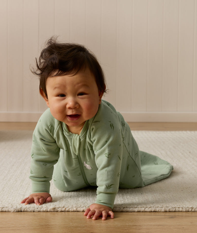 A baby wearing a long-sleeved, sage green leaf-patterned sleeping bag crawls on a light rug in front of a neutral wood-paneled wall, smiling toward the camera.