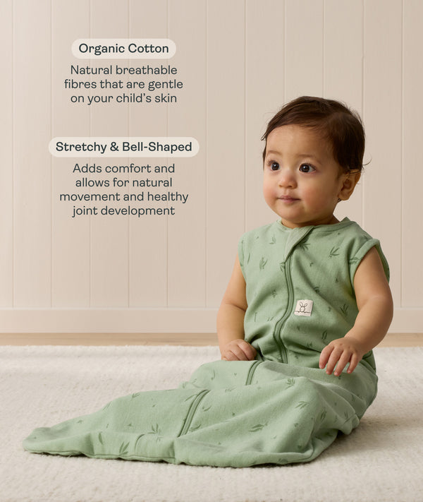 Infant sitting on a white mat, wearing a green Sleeping Bag 1.0 TOG with text highlighting organic cotton and stretchy & bell-shaped design.