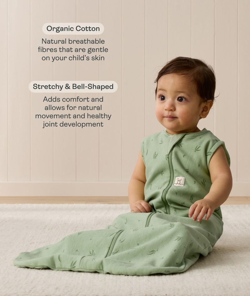 Infant sitting on a white mat, wearing a green Sleeping Bag 1.0 TOG with text highlighting organic cotton and stretchy & bell-shaped design.