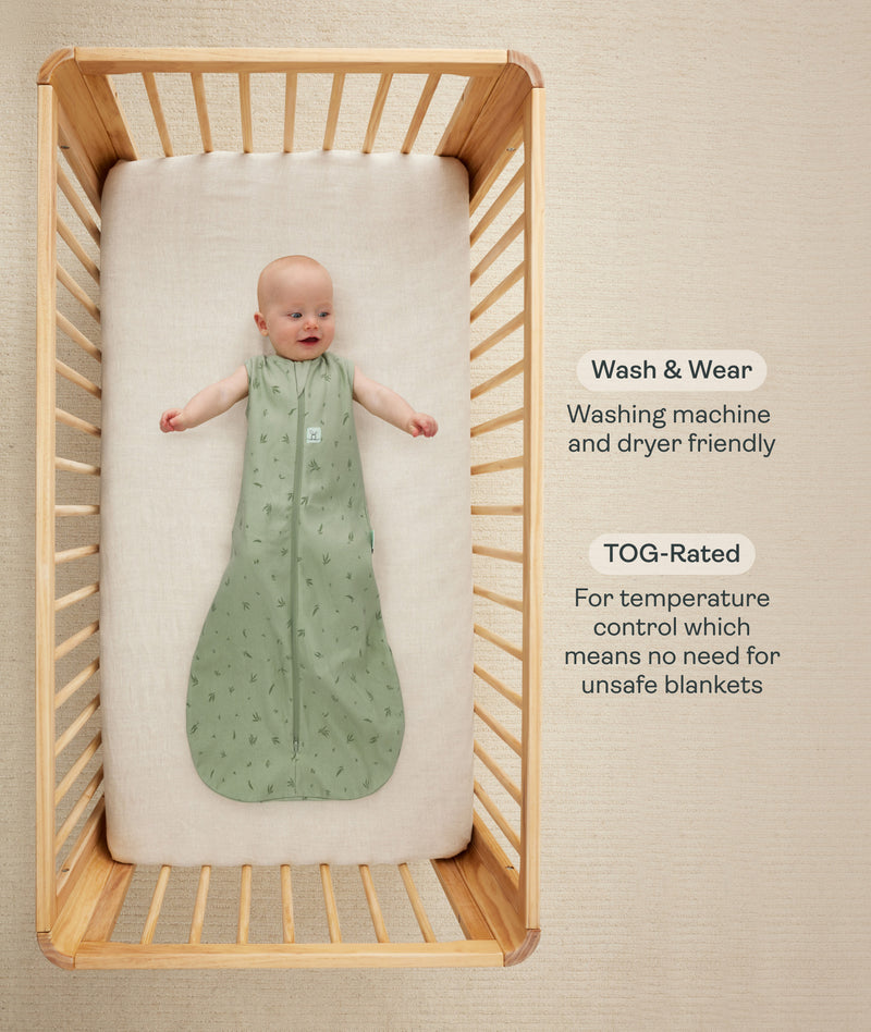 Baby lying on their back inside a wooden cot wearing a green sleeveless Sleeping Bag 1.0 TOG with text callouts highlighting wash & wear care and TOG rating features.