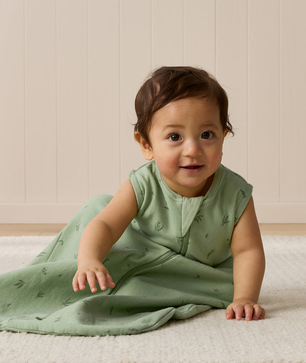 A baby wearing a sleeveless, sage green leaf-patterned sleeping bag sits on a light rug, leaning forward with one hand resting on the floor.