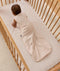 An overhead view of a baby wearing a pattern-printed sleeveless Sleeping Bag, crawling on their hands and knees inside a wooden cot with a grid sheet.