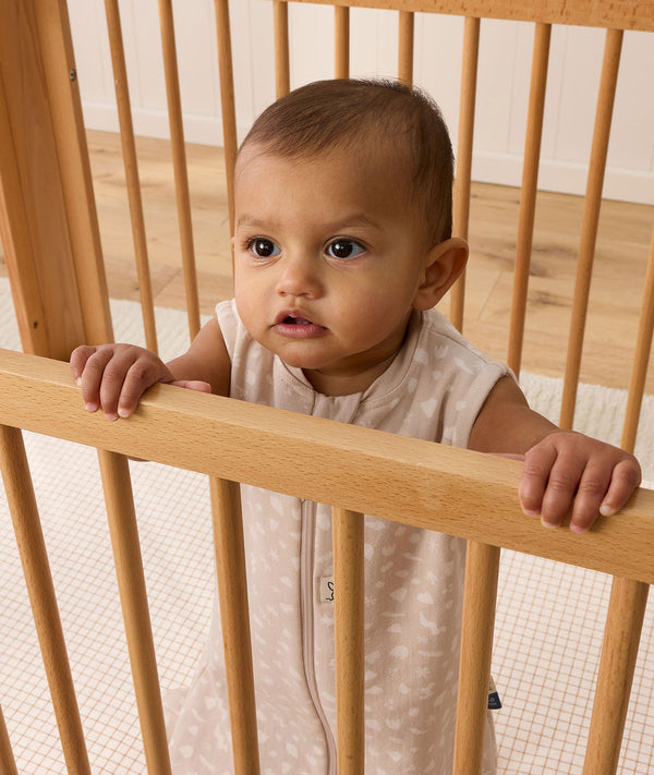 A baby wearing a printed sleeveless Sleeping Bag, standing and holding onto the rail of a wooden cot.