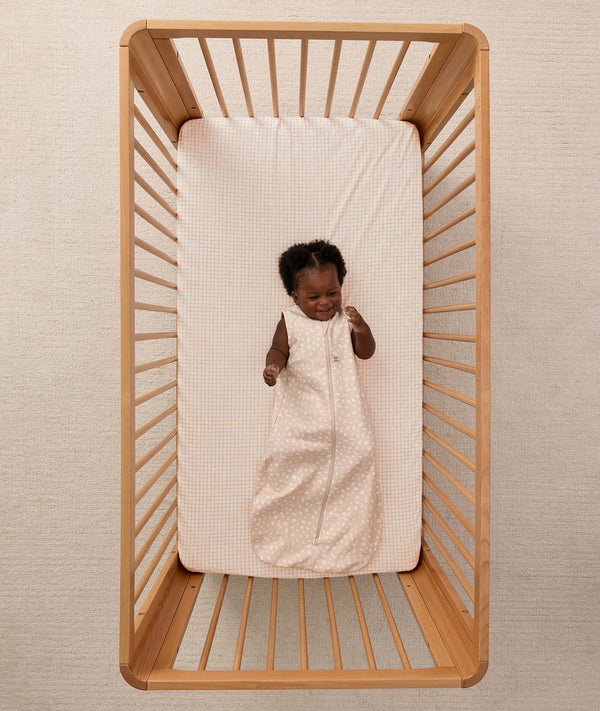A smiling baby lying inside a wooden cot, wearing a sleeveless ergoPouch Sleeping Bag 1.0 TOG