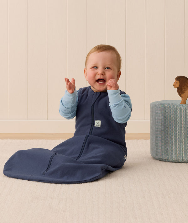 A happy baby sitting upright is wearing a navy blue sleeping bag and a long sleeve layer underneath.