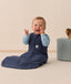 A happy baby sitting upright is wearing a navy blue sleeping bag and a long sleeve layer underneath.