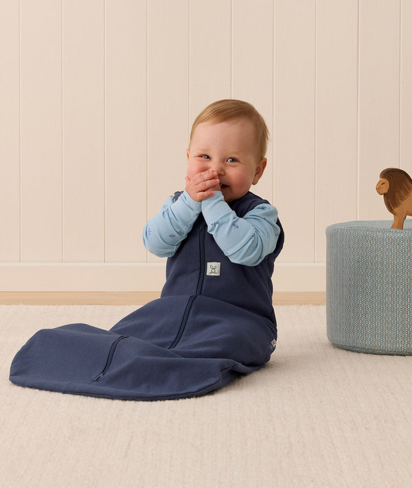 Baby sitting upright wearing a navy blue sleeping bag over a long sleeve layer.