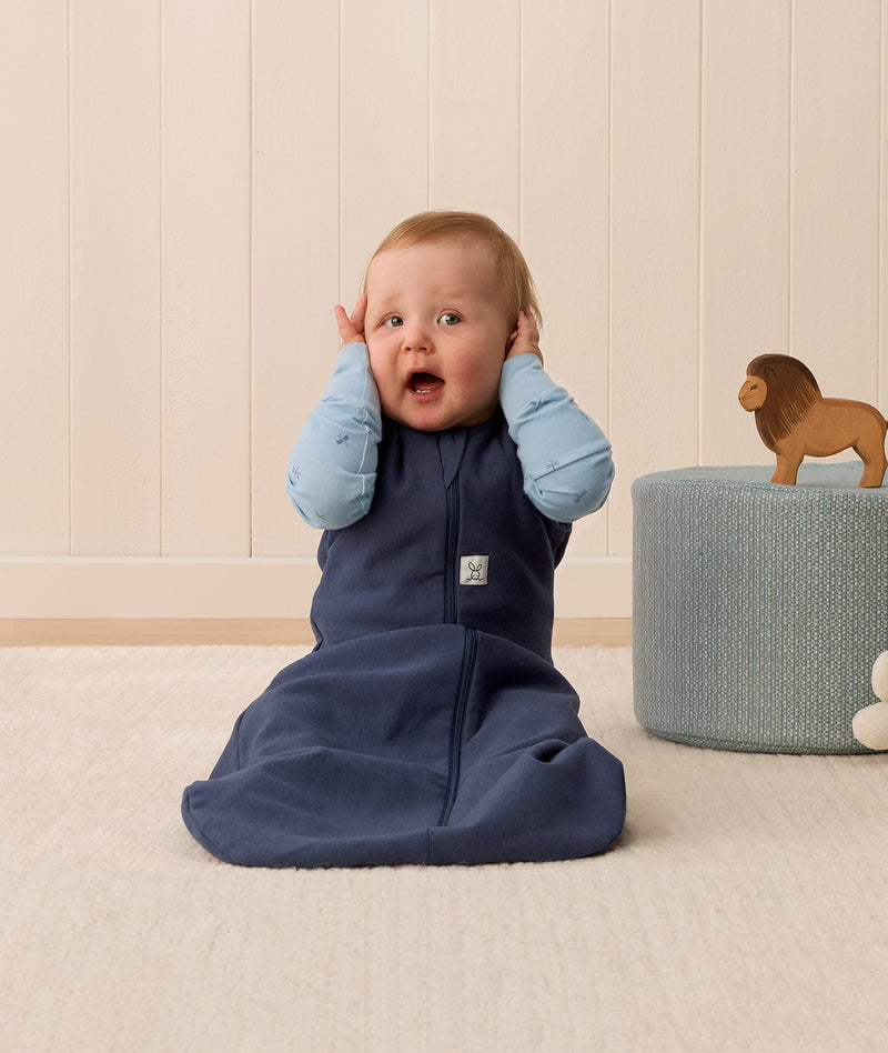 Baby sitting upright wearing a navy blue sleeping bag, featuring a zip-front design and soft, breathable fabric.