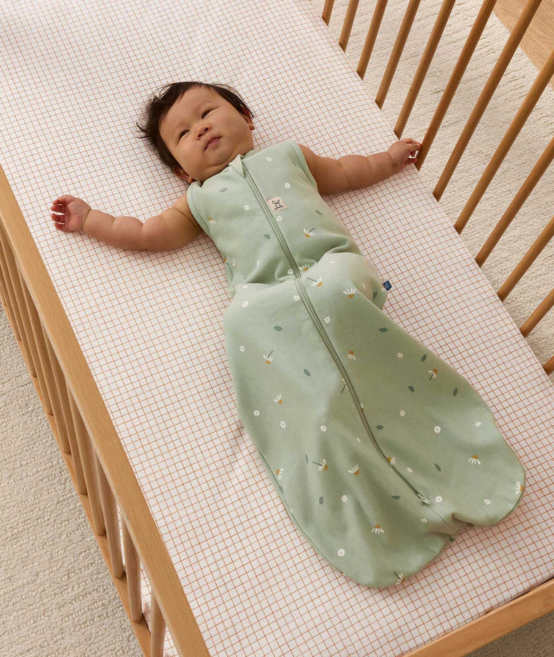 Infant lying in a wooden cot with a grid-patterned sheet, wearing a sage green sleeveless Sleeping Bag with floral pattern.