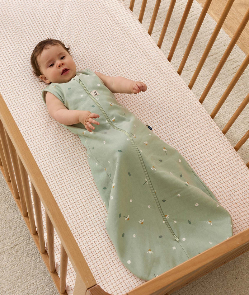 Infant dressed in a sleeveless ergoPouch Sleeping Bag 1.0 TOG, lying in a wooden cot with a grid-patterned sheet