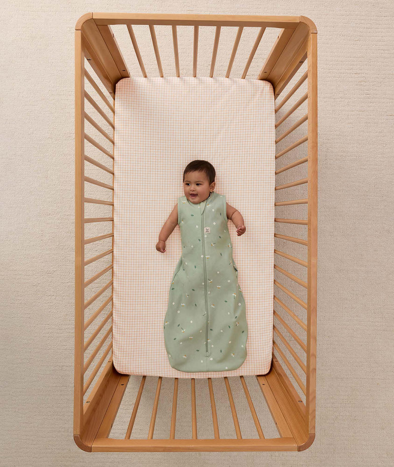 An overhead shot of an infant lying in a wooden cot, wearing a sleeveless floral-patterned Sleeping Bag 1.0 TOG