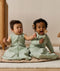 Two infants are sitting side-by-side while holding a wooden toy. One is wearing a sleeveless Sleeping Bag, while the other is wearing the same print with sleeves.