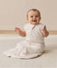 Smiling infant sitting on a white mat, wearing a sleeveless Sleeping Bag in Oatmeal Marle
