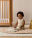 A smiling infant sitting on a beige quilted rug in a nursery while wearing a sleeveless, animal-patterned Sleeping Bag next to a wooden cot.
