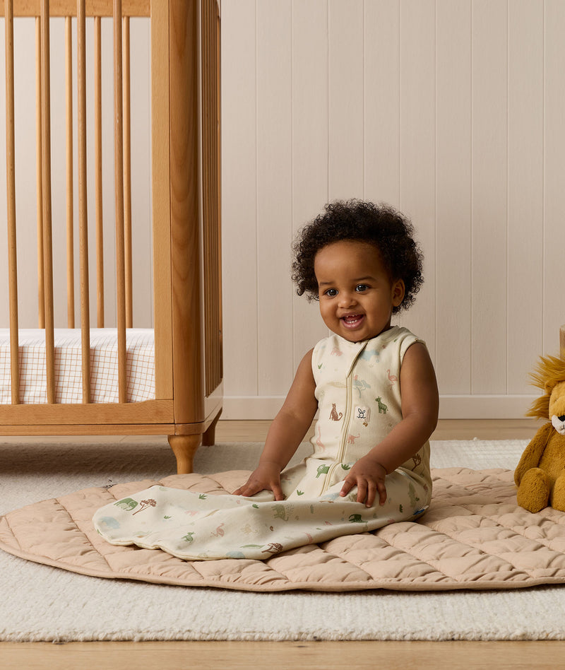 A smiling infant sitting on a beige quilted rug in a nursery while wearing a sleeveless, animal-patterned Sleeping Bag next to a wooden cot.