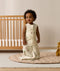 An infant standing upright on a beige quilted mat in a nursery while wearing an animal-patterned sleeveless Sleeping Bag next to a wooden cot.