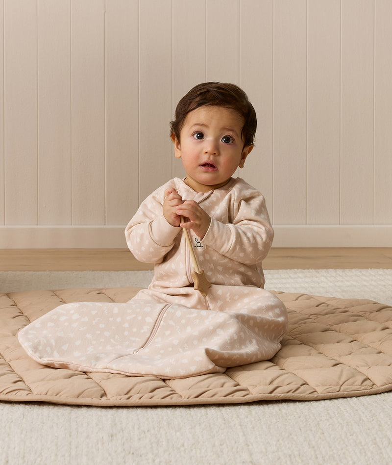 Infant sitting on a quilted mat while holding a wooden toy, wearing a long-sleeve patterned Sleeping Bag 1.0 TOG