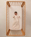 An overhead view on an infant lying in a wooden cot with a grid-patterned sheet, wearing a long sleeve Sleeping Bag 1.0 TOG