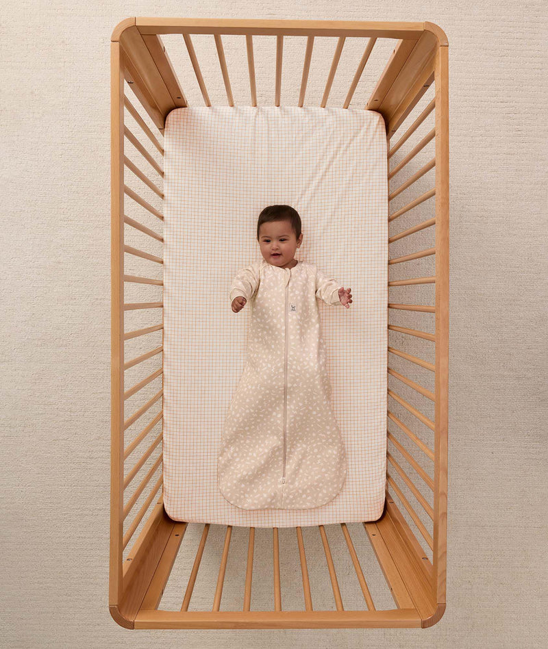 An overhead view on an infant lying in a wooden cot with a grid-patterned sheet, wearing a long sleeve Sleeping Bag 1.0 TOG