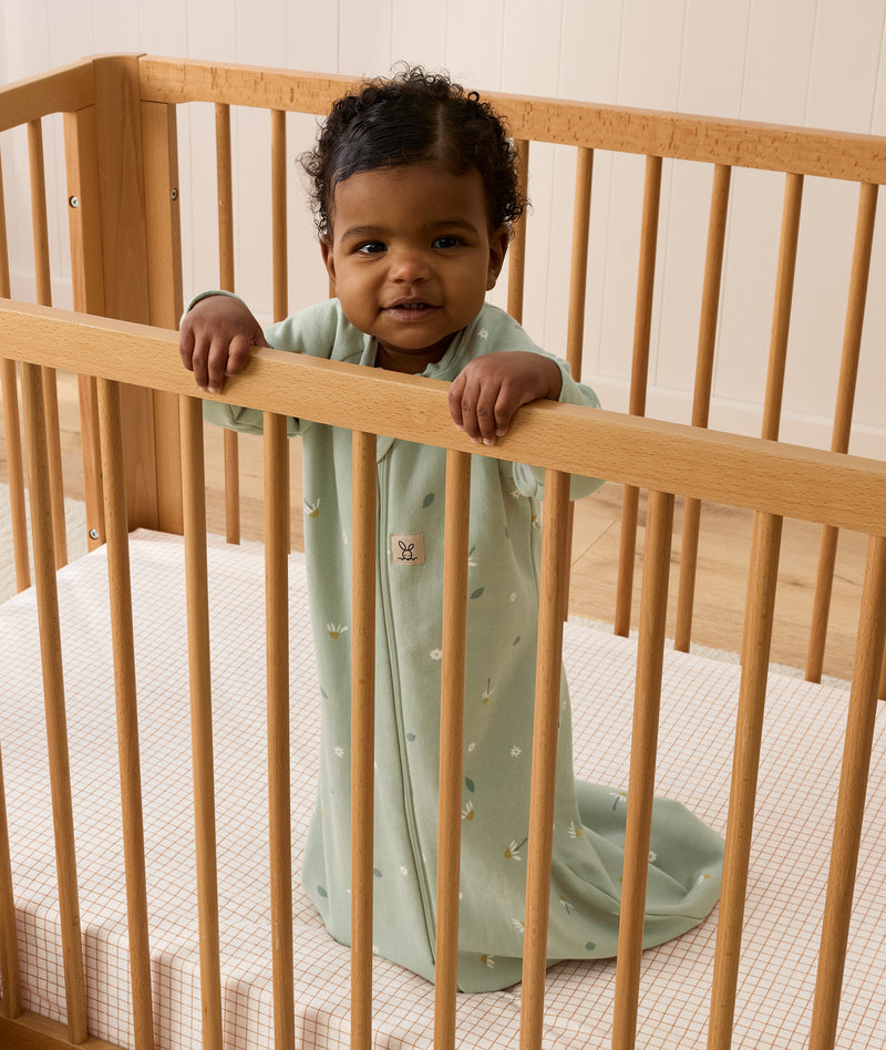 Infant wearing a sage green long-sleeve Sleeping Bag 1.0 TOG, standing inside a wooden cot