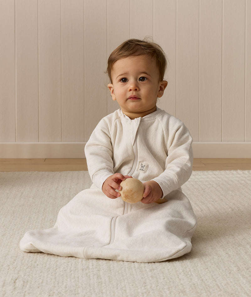 Infant sitting, wearing a sleeved Sleeping Bag 1.0 TOG Oatmeal Marle, while holding a wooden toy.