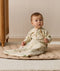 A toddler sitting on a padded mat wearing a beige safari-patterned Sleeping Bag with sleeves and holding a small wooden wand.