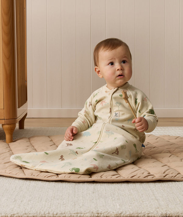 A toddler sitting on a padded mat wearing a beige safari-patterned Sleeping Bag with sleeves and holding a small wooden wand.