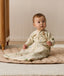 A toddler sitting on a padded mat wearing a beige safari-patterned Sleeping Bag with sleeves and holding a small wooden wand.