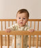 A toddler standing inside a wooden cot, holding onto the top rail while wearing a safari-printed sleeping bag.