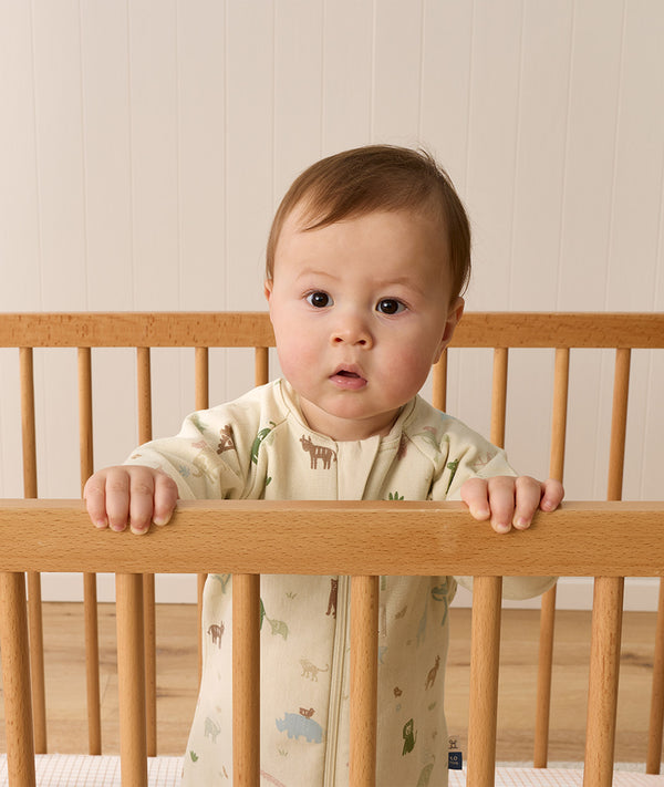 A toddler standing inside a wooden cot, holding onto the top rail while wearing a safari-printed sleeping bag.
