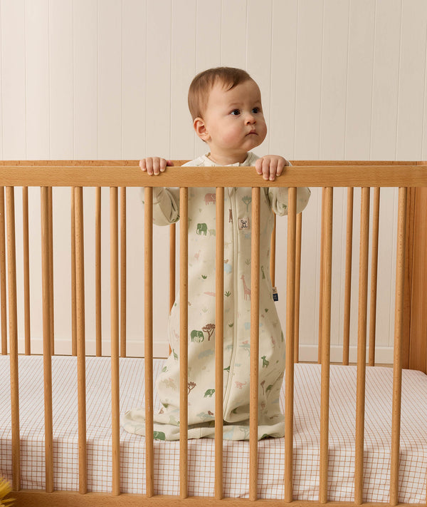 Infant standing inside a wooden cot wearing a long-sleeve sleeping bag
