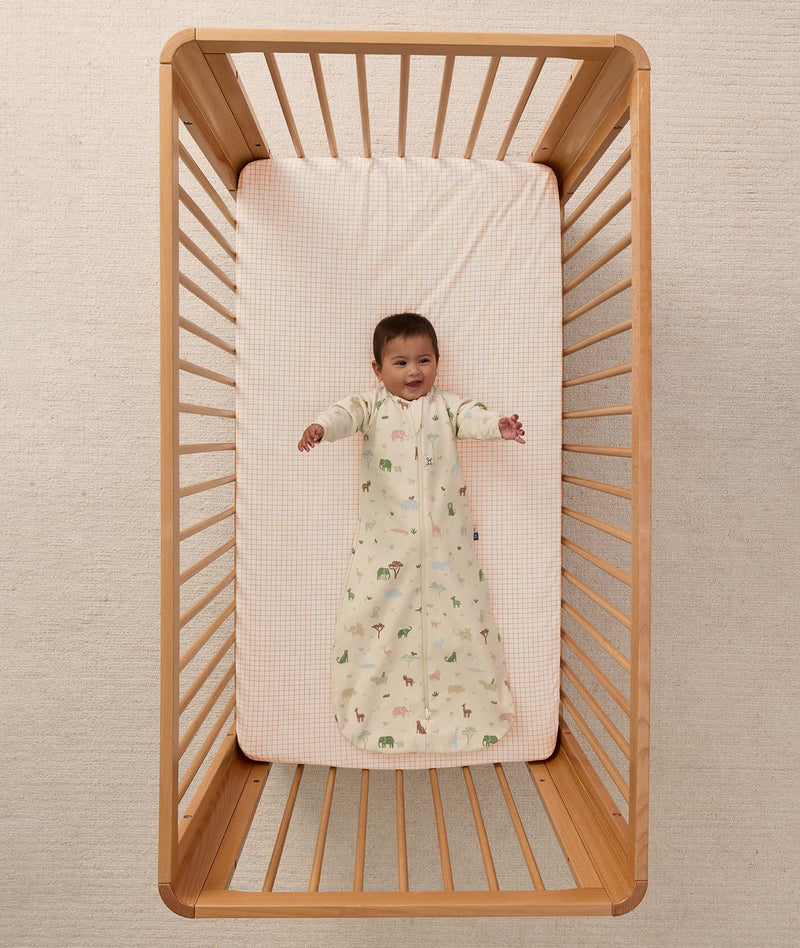 Overhead view of a baby lying on their back in a wooden cot, wearing a safari-patterned long-sleeve Sleeping Bag with arms outstretched.