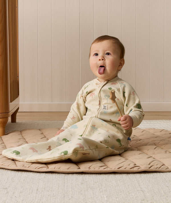 A playful baby sitting on a padded mat sticking out their tongue, holding a wooden star wand while wearing a safari-patterned sleeping bag.