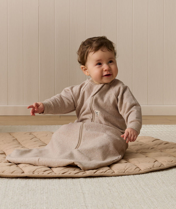 Infant wearing a long-sleeve Sleeping Bag 1.0 TOG Taupe Marle, sitting on a quilted mat