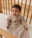 A baby wearing a long-sleeved beige Sleeping Bag 1.0 TOG, sitting upright inside a wooden cot with a grid-patterned sheet.
