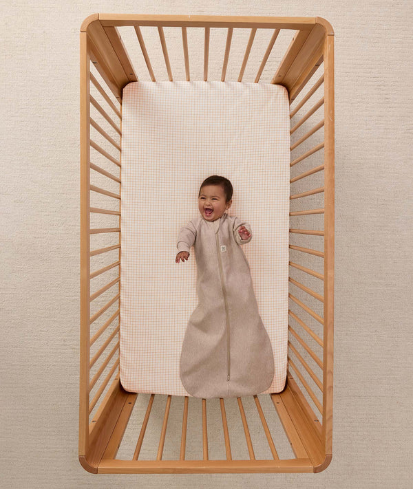 A happy baby lying in a wooden cot, wearing a sleeved Sleeping Bag 1.0 TOG Taupe Marle